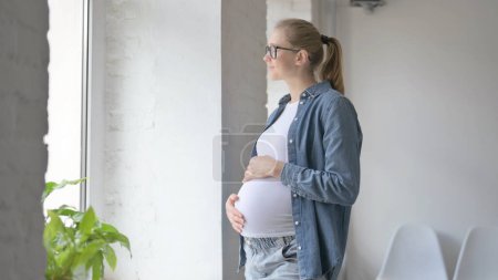 Foto de Embarazada hermosa mujer sosteniendo su gran barriga mientras mira fuera de ventana - Imagen libre de derechos