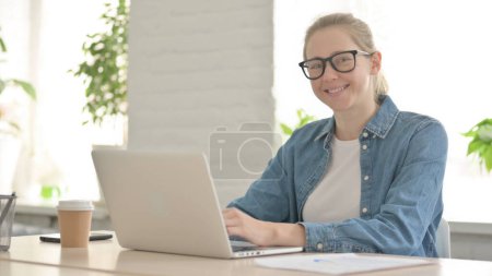Téléchargez les photos : Belle femme souriant à la caméra tout en utilisant un ordinateur portable dans le bureau - en image libre de droit