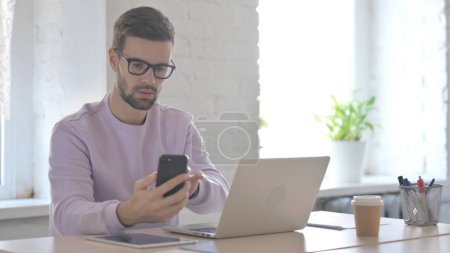Photo for Young Adult Man using Smartphone while using Laptop - Royalty Free Image