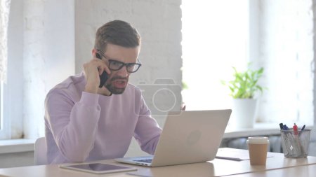 Foto de Hombre joven adulto enojado hablando por teléfono mientras trabaja en el ordenador portátil - Imagen libre de derechos