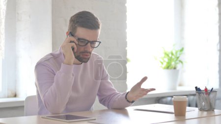 Téléchargez les photos : Agacé jeune homme adulte parlant avec colère sur Smartphone au bureau - en image libre de droit