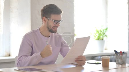 Téléchargez les photos : Jeune homme adulte excité lisant des documents et applaudissant au bureau - en image libre de droit