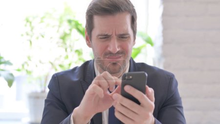 Photo for The Upset Young Adult Businessman Reacting to Loss on Smartphone in Office - Royalty Free Image