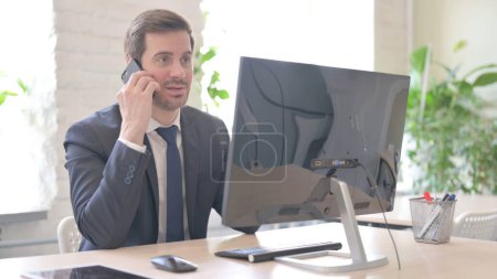 Foto de The Young Adult Businessman Talking on Phone while Working on Computer - Imagen libre de derechos