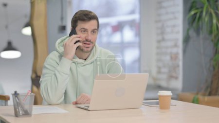 Foto de Hombre adulto joven hablando por teléfono mientras usa el ordenador portátil - Imagen libre de derechos