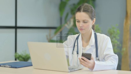 Téléchargez les photos : Jeune femme médecin en utilisant Smartphone tout en utilisant ordinateur portable - en image libre de droit