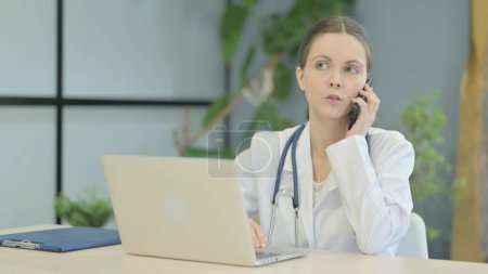 Foto de Doctora joven hablando por teléfono mientras usa el ordenador portátil - Imagen libre de derechos
