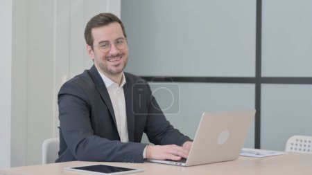 Foto de Empresario sonriendo a la cámara mientras usa el ordenador portátil - Imagen libre de derechos