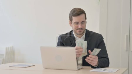 Téléchargez les photos : Homme d'affaires compte dollar tout en travaillant sur ordinateur portable - en image libre de droit