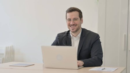 Téléchargez les photos : Homme d'affaires sourire à la caméra tout en utilisant un ordinateur portable - en image libre de droit