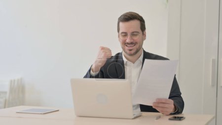 Téléchargez les photos : Homme d'affaires Célébrant le succès tout en travaillant sur ordinateur portable et documents - en image libre de droit