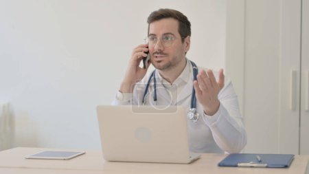 Foto de Doctor hablando por teléfono mientras usa el portátil - Imagen libre de derechos