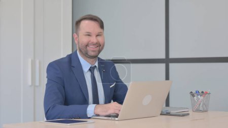 Téléchargez les photos : Homme d'affaires souriant à la caméra tout en travaillant sur ordinateur portable - en image libre de droit