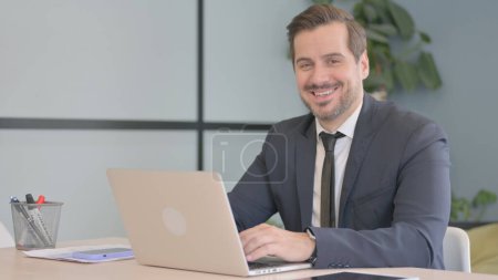 Téléchargez les photos : Homme d'affaires souriant à la caméra tout en travaillant sur ordinateur portable - en image libre de droit