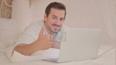 Téléchargez les photos : Jeune homme faisant pouce vers le haut tout en utilisant l'ordinateur portable allongé sur l'estomac - en image libre de droit