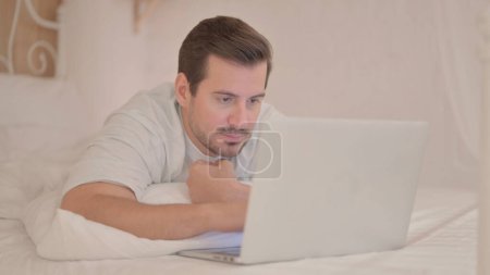 Téléchargez les photos : Jeune homme travaillant sur ordinateur portable tout en s'allongeant sur l'estomac au lit - en image libre de droit