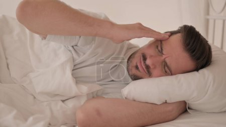 Téléchargez les photos : Jeune homme avec mal de tête couché au lit sur le côté - en image libre de droit