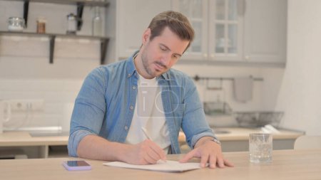 Téléchargez les photos : Jeune homme écrivant tout en faisant de la paperasserie - en image libre de droit
