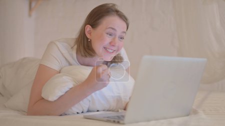 Foto de Mujer joven celebrando en el ordenador portátil mientras se acuesta en la cama con el estómago - Imagen libre de derechos