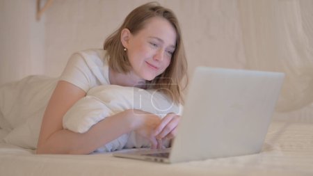 Téléchargez les photos : Chat vidéo en ligne par Jeune femme sur ordinateur portable tout en s'allongeant sur l'estomac au lit - en image libre de droit