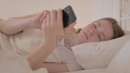 Téléchargez les photos : Vue latérale de la jeune femme à l'aide d'un téléphone intelligent au lit - en image libre de droit