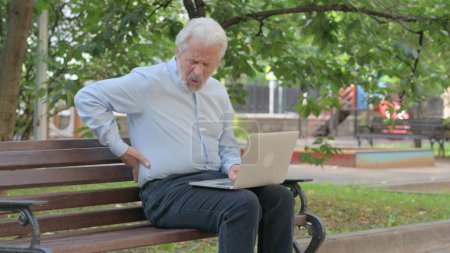 Téléchargez les photos : Vieil homme âgé utilisant un ordinateur portable avec douleur au dos tout en étant assis à l'extérieur - en image libre de droit