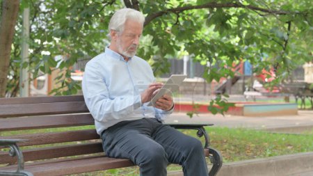 Téléchargez les photos : Vieil homme âgé utilisant la tablette tout en étant assis en plein air sur un banc - en image libre de droit