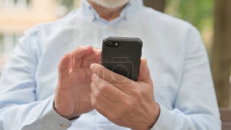 Foto de Primer plano del anciano mayor navegando por el teléfono inteligente mientras está sentado al aire libre en un banco - Imagen libre de derechos