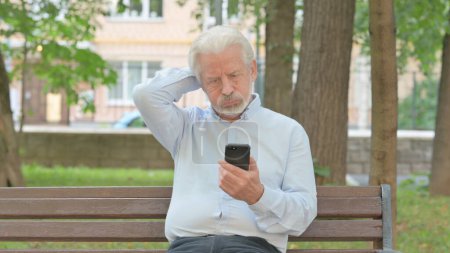 Téléchargez les photos : Triste vieil homme âgé avec perte en ligne sur Smartphone tout en étant assis sur un banc - en image libre de droit