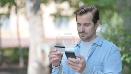 Photo for Excited Young Man Shopping Online via phone while Sitting Outdoor on a Bench - Royalty Free Image