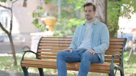 Téléchargez les photos : Sérieux jeune homme assis en plein air sur le banc - en image libre de droit
