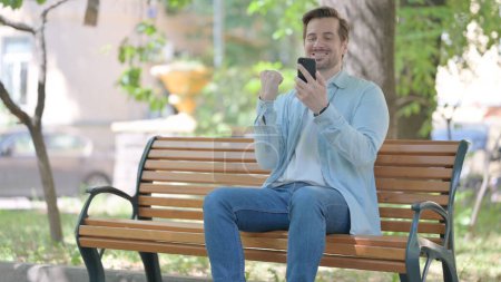 Téléchargez les photos : Jeune homme célébrant le succès sur Smartphone tout en étant assis à l'extérieur sur un banc - en image libre de droit