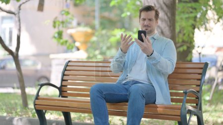Foto de Joven sorprendido por la pérdida en el teléfono inteligente mientras está sentado al aire libre en un banco - Imagen libre de derechos