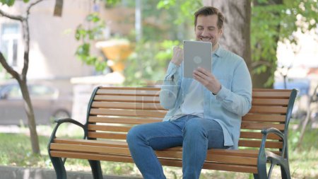 Téléchargez les photos : Jeune homme célébrant le succès sur tablette tout en étant assis à l'extérieur sur un banc - en image libre de droit
