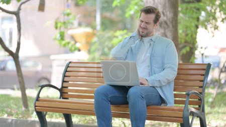 Téléchargez les photos : Jeune homme avec douleur au cou travaillant sur ordinateur portable en plein air - en image libre de droit
