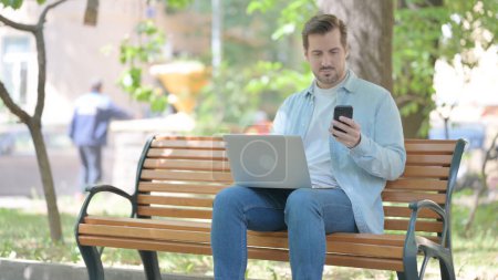 Téléchargez les photos : Jeune homme utilisant Smartphone et ordinateur portable tout en étant assis en plein air - en image libre de droit