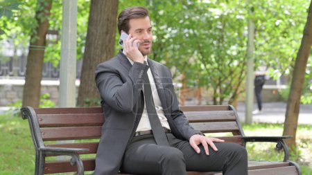 Téléchargez les photos : Jeune homme d'affaires Parler au téléphone tout en étant assis à l'extérieur sur un banc - en image libre de droit