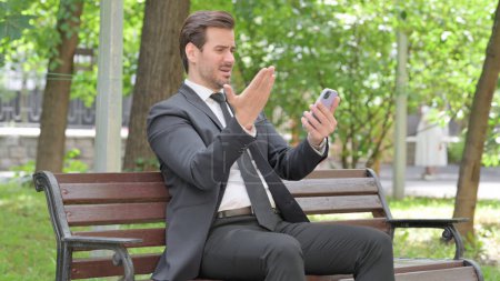 Téléchargez les photos : Jeune homme d'affaires choqué par la perte sur Smartphone tout en étant assis à l'extérieur sur un banc - en image libre de droit