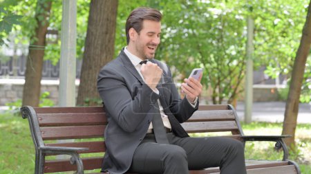 Téléchargez les photos : Jeune homme d'affaires célébrant le succès sur Smartphone assis à l'extérieur sur un banc - en image libre de droit