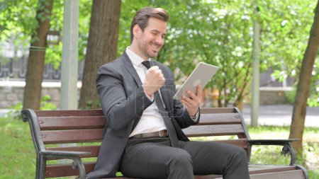 Téléchargez les photos : Jeune homme d'affaires célébrant le succès sur tablette tout en étant assis à l'extérieur sur un banc - en image libre de droit