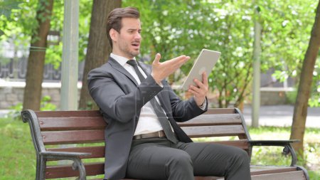 Téléchargez les photos : Jeune homme d'affaires bouleversé par la perte sur tablette tout en étant assis à l'extérieur sur un banc - en image libre de droit