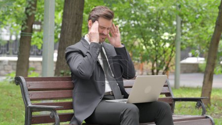 Téléchargez les photos : Jeune homme d'affaires avec maux de tête travaillant sur ordinateur portable en plein air - en image libre de droit