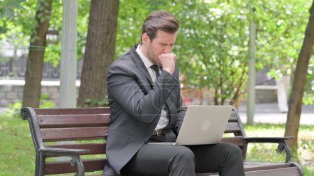 Téléchargez les photos : Toux jeune homme d'affaires utilisant un ordinateur portable en plein air - en image libre de droit