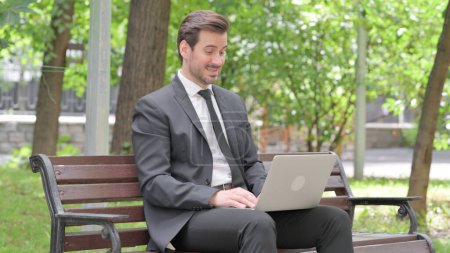 Téléchargez les photos : Chat vidéo en ligne par jeune homme d'affaires sur ordinateur portable en plein air - en image libre de droit