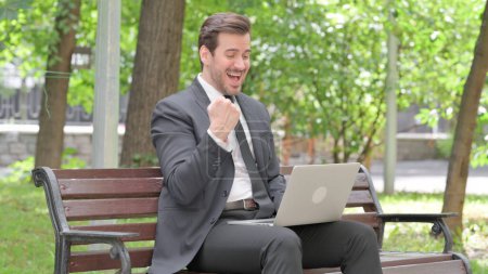 Téléchargez les photos : Jeune homme d'affaires excité célébrant le succès sur ordinateur portable en plein air - en image libre de droit