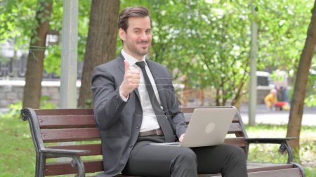 Téléchargez les photos : Pouces levées par jeune homme d'affaires sur ordinateur portable en plein air - en image libre de droit