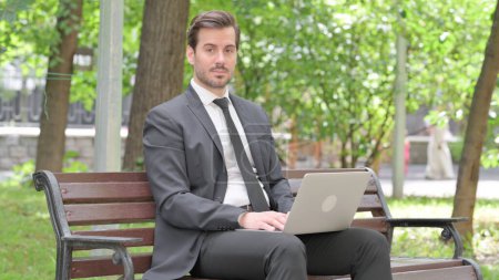 Photo for Young Businessman Looking at Camera while Working on Laptop Outdoor - Royalty Free Image