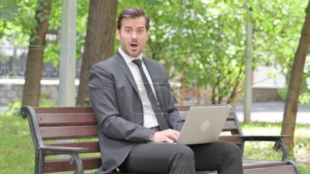 Téléchargez les photos : Choqué jeune homme d'affaires regardant la caméra tout en travaillant sur ordinateur portable en plein air - en image libre de droit