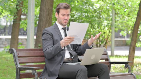 Téléchargez les photos : Jeune homme d'affaires se sentant bouleversé tout en lisant contrat et en utilisant ordinateur portable en plein air - en image libre de droit