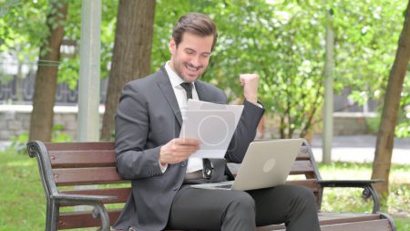 Téléchargez les photos : Jeune homme d'affaires Célébration tout en travaillant sur ordinateur portable et documents en plein air - en image libre de droit